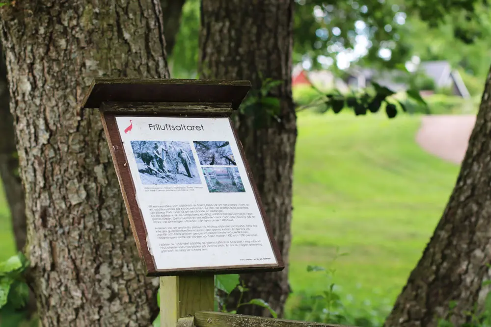 En skylt av trä utomhus i parkmiljö. Skylten beskriver friluftsaltaret som en gång fanns i byn.