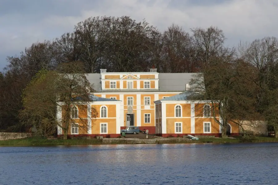 1800-tals slott i gult med vita detaljer, med två fristående kvadratiska byggnader framför i samma färg och stil.