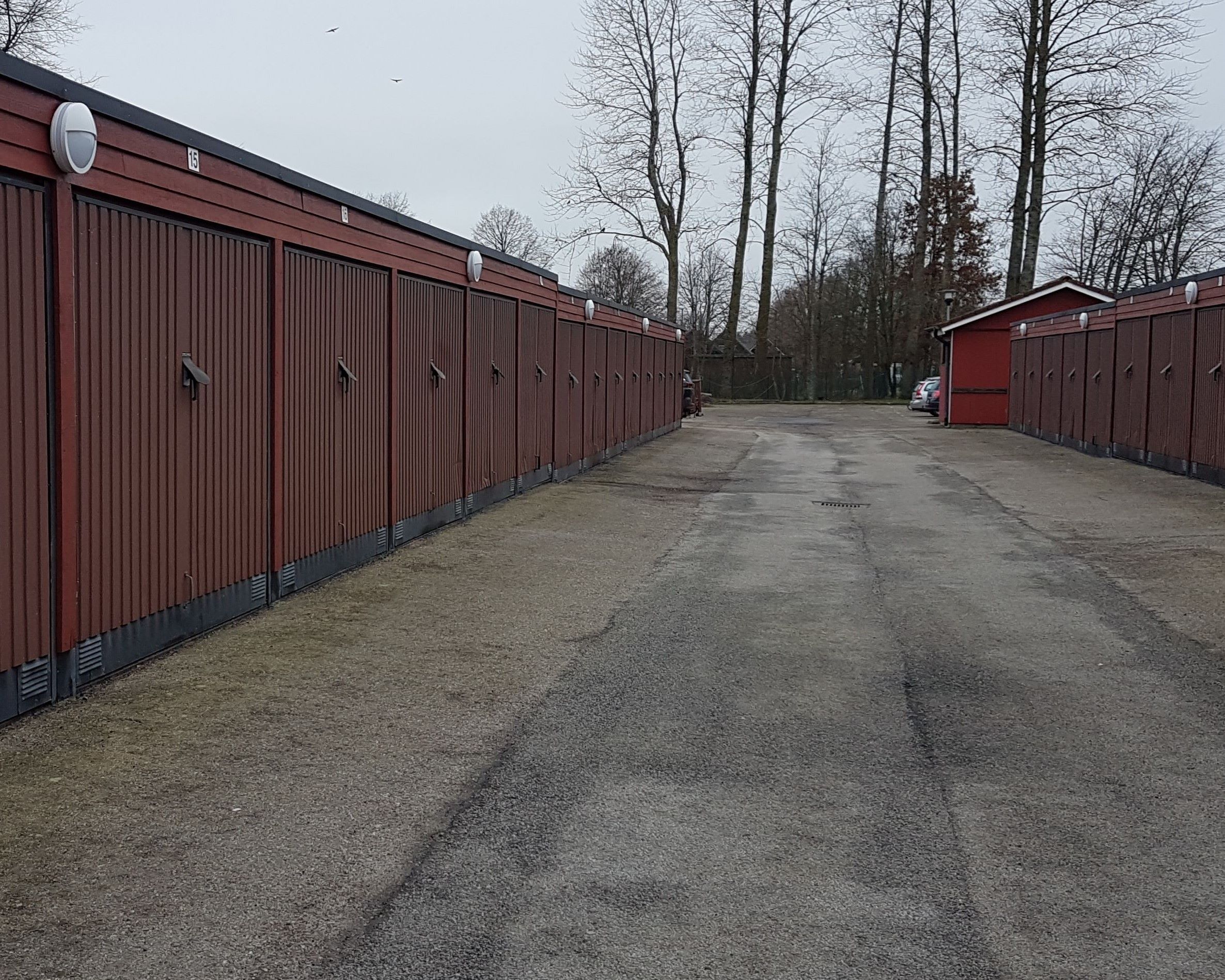 Röda garagelängor med brundröda portar som står mittemott varandra. Asfalterad väd mellan och träd längst bort vid vändzonen.
