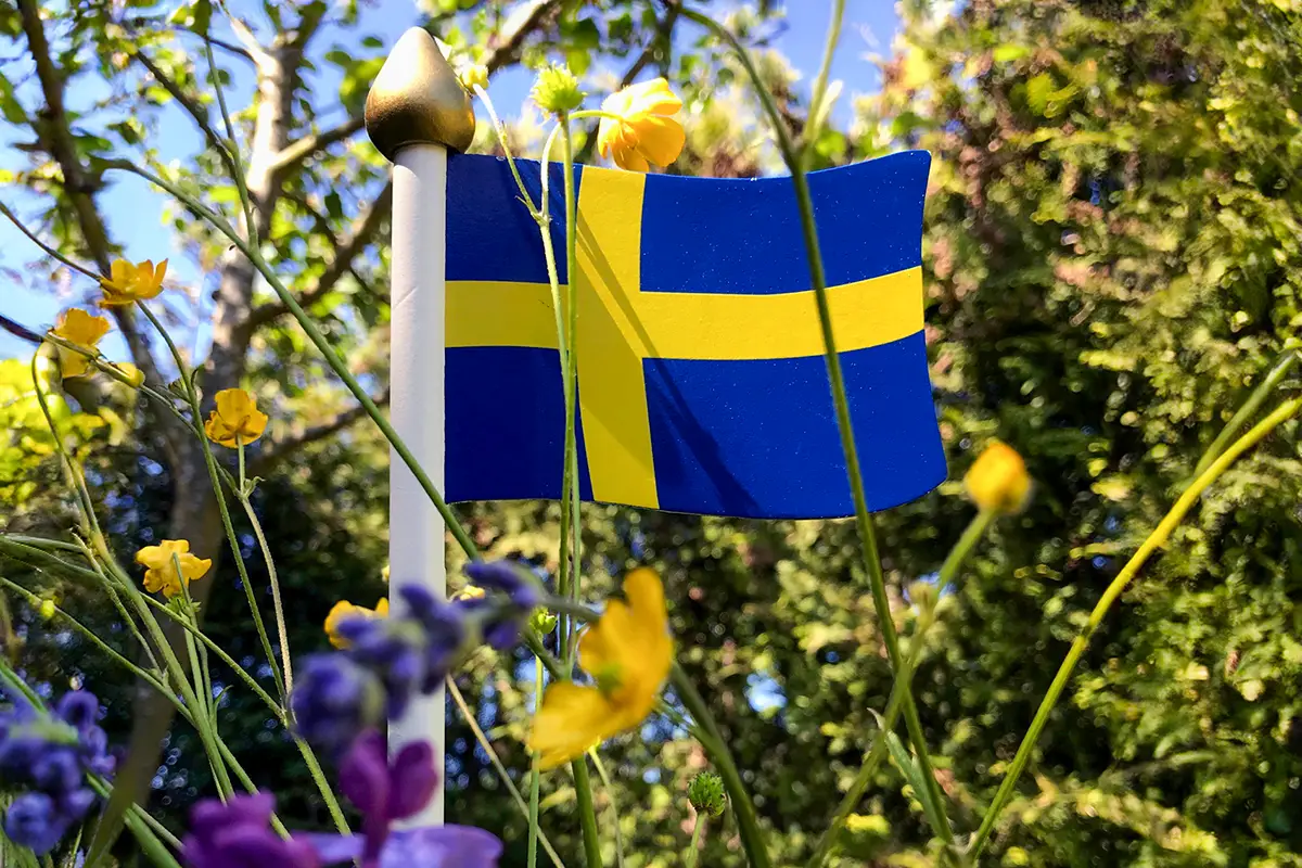 Liten svensk flagga i trä med ängsblommor runt omkring.