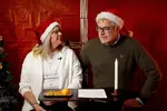 Britt-Marie och Per-Martin iklädda tomteluvor, sittandes framför en bakgrund inpackad i rött julklappspapper med skimrande snören. Sidan om står en julgran. 